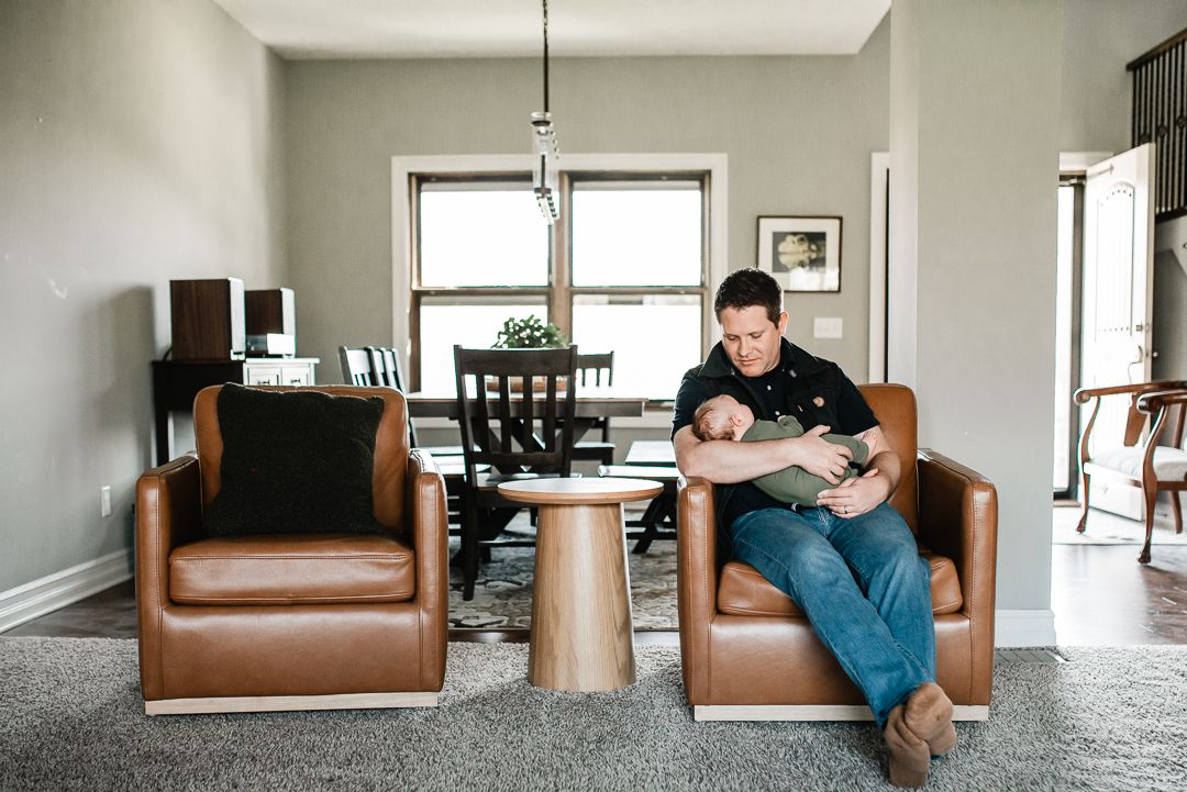 A father sits in a brown leather chair holding his newborn baby in a modern living room with neutral decor.