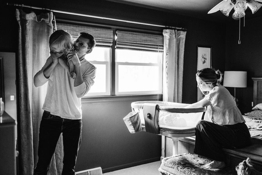 Black and white photo of a man holding a baby near a window while a woman makes the bed in a cozy bedroom.