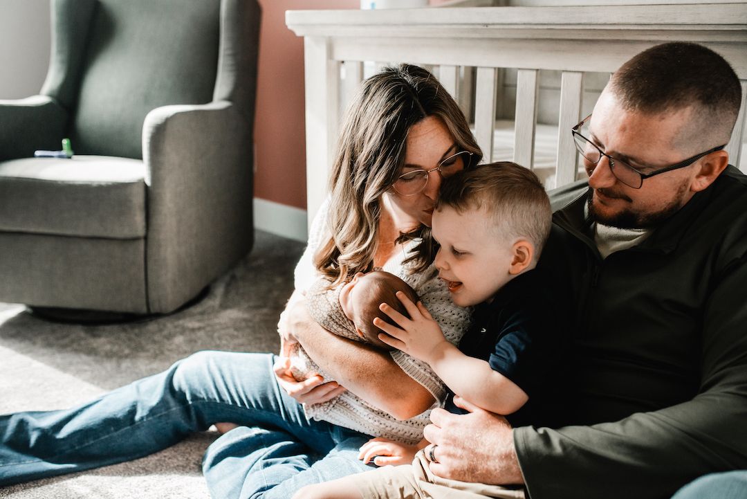 A young family sits on the floor together, parents lovingly embracing their toddler son in a cozy nursery room.