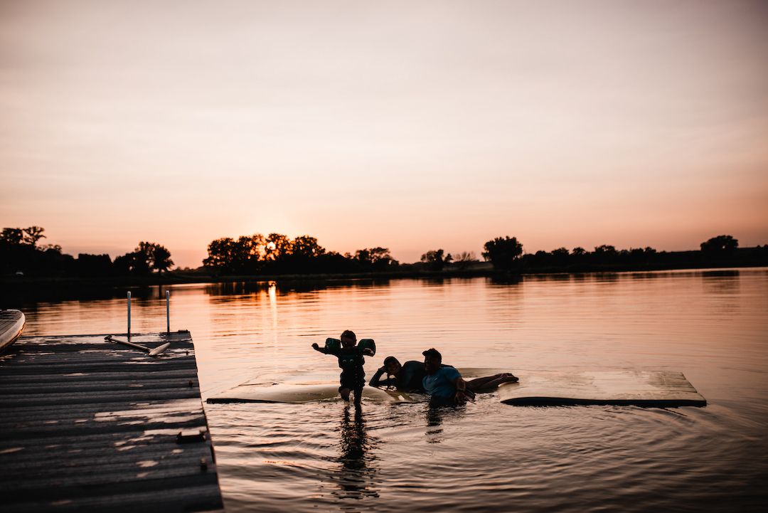 Silhouettes of people swimming and playing on a dock at sunset over a calm lake.