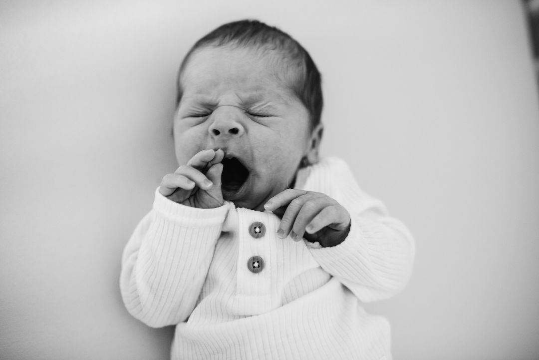 Black and white photo of a newborn baby yawning with hand near mouth, wearing a white onesie.