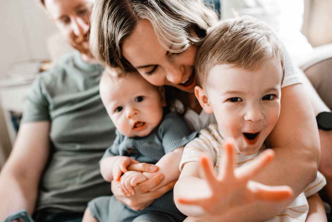 Mother kissing baby while toddler reaches toward camera in a cozy indoor family moment.