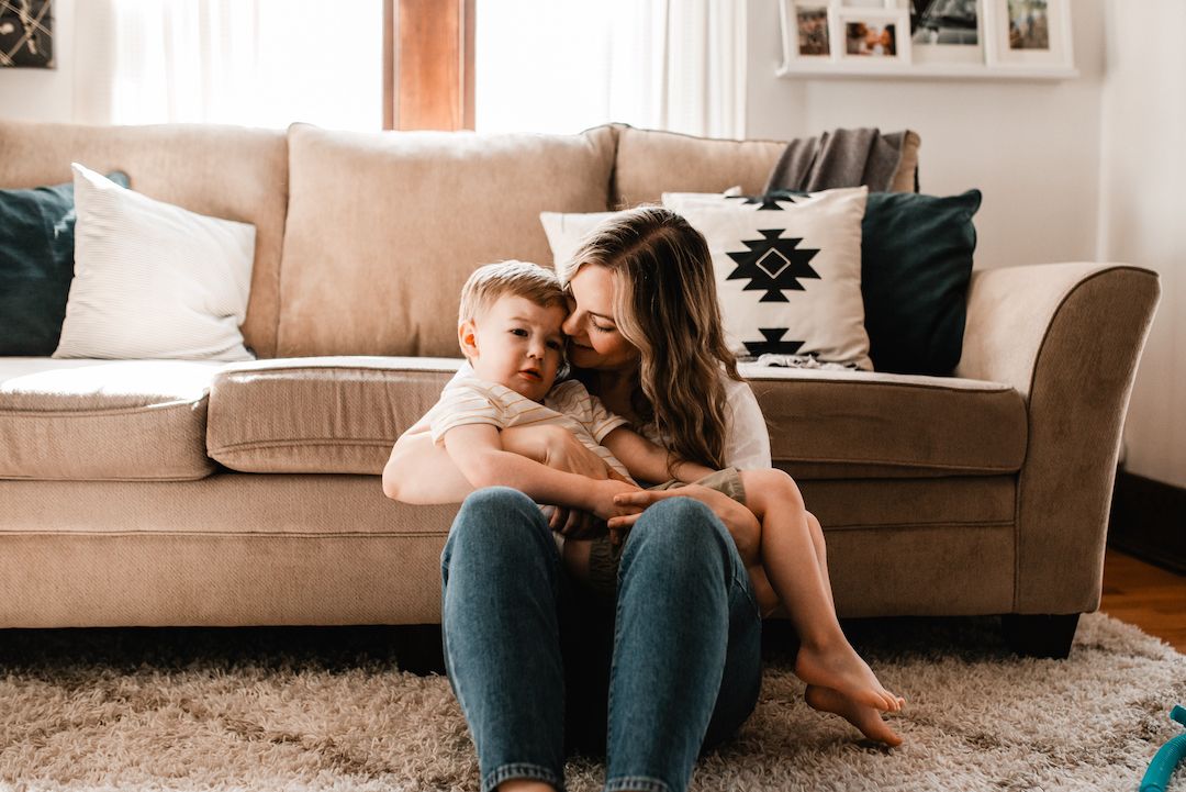 Mother sitting on a rug hugging her young son in front of a beige sofa.