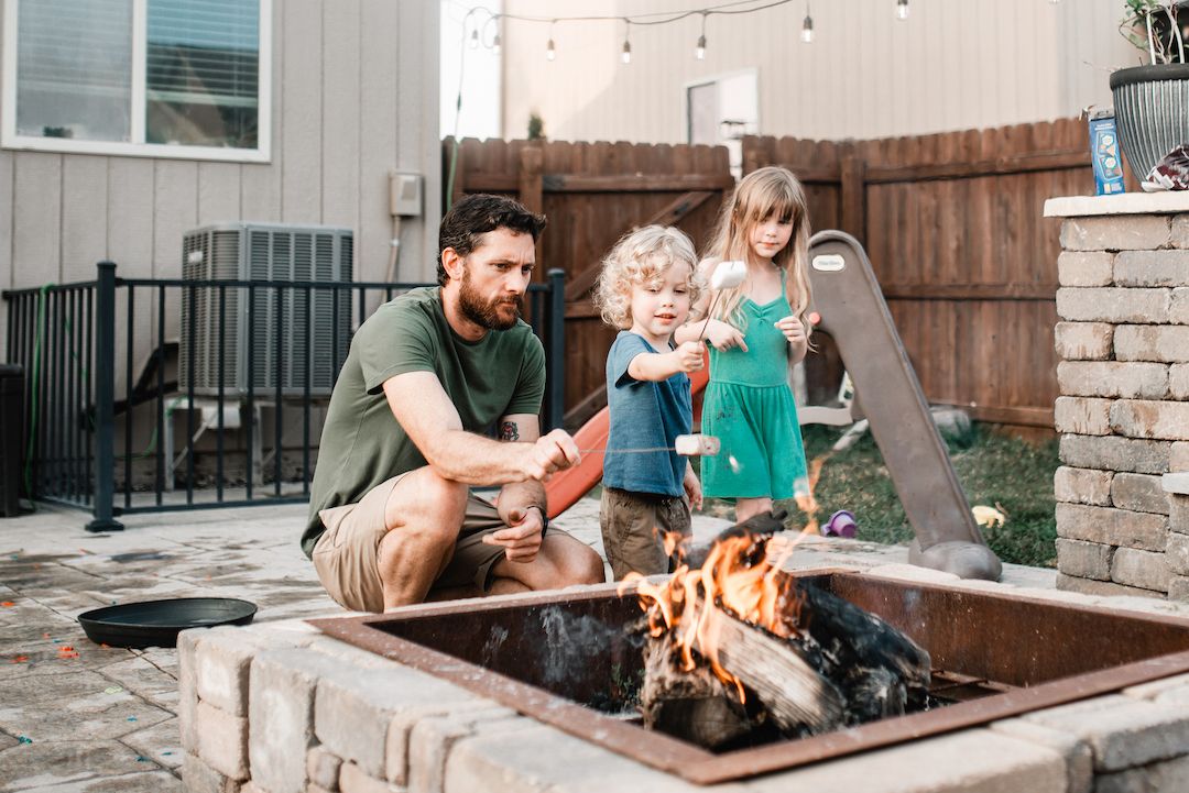 Father and two children roasting marshmallows over backyard fire pit.