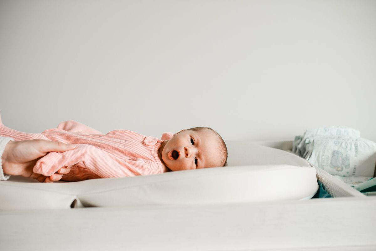 Newborn baby in pink onesie lying on white changing pad with mouth open, diaper visible in background.