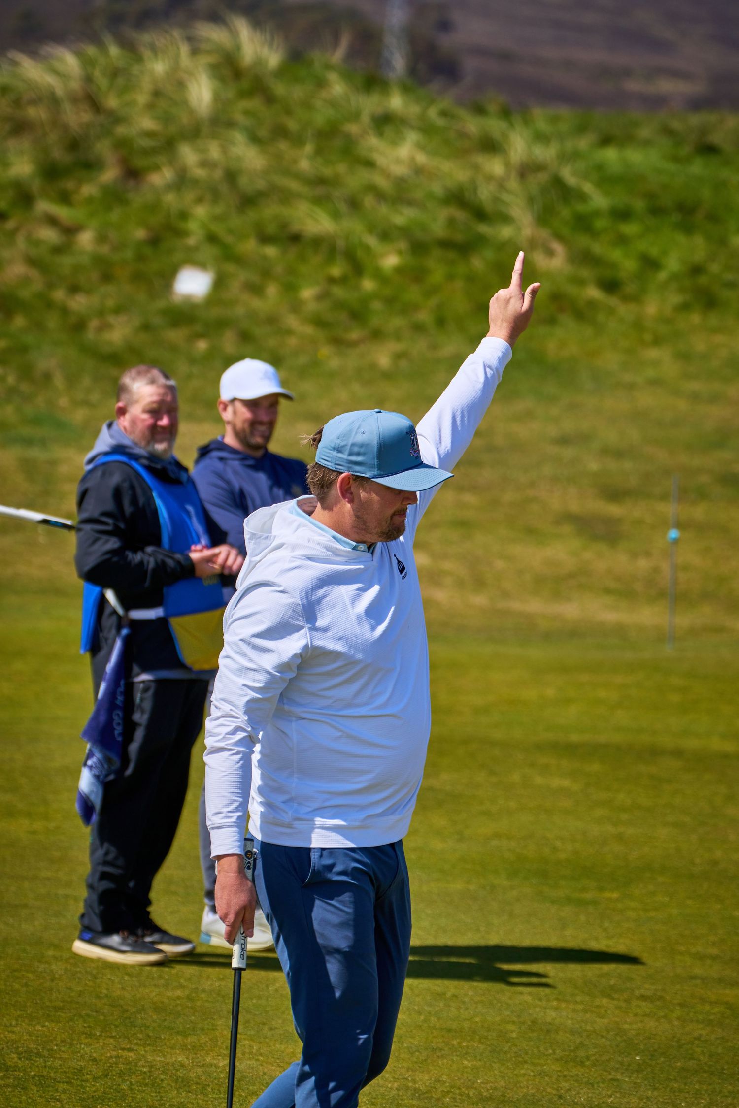 Golfer raises hand in victory on green course, with caddie and fellow player watching in background.