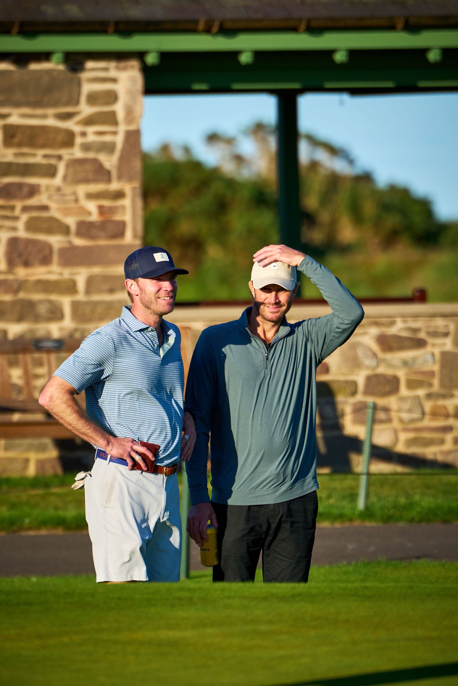Two male golfers in caps chat on a sunny golf course near a stone clubhouse.