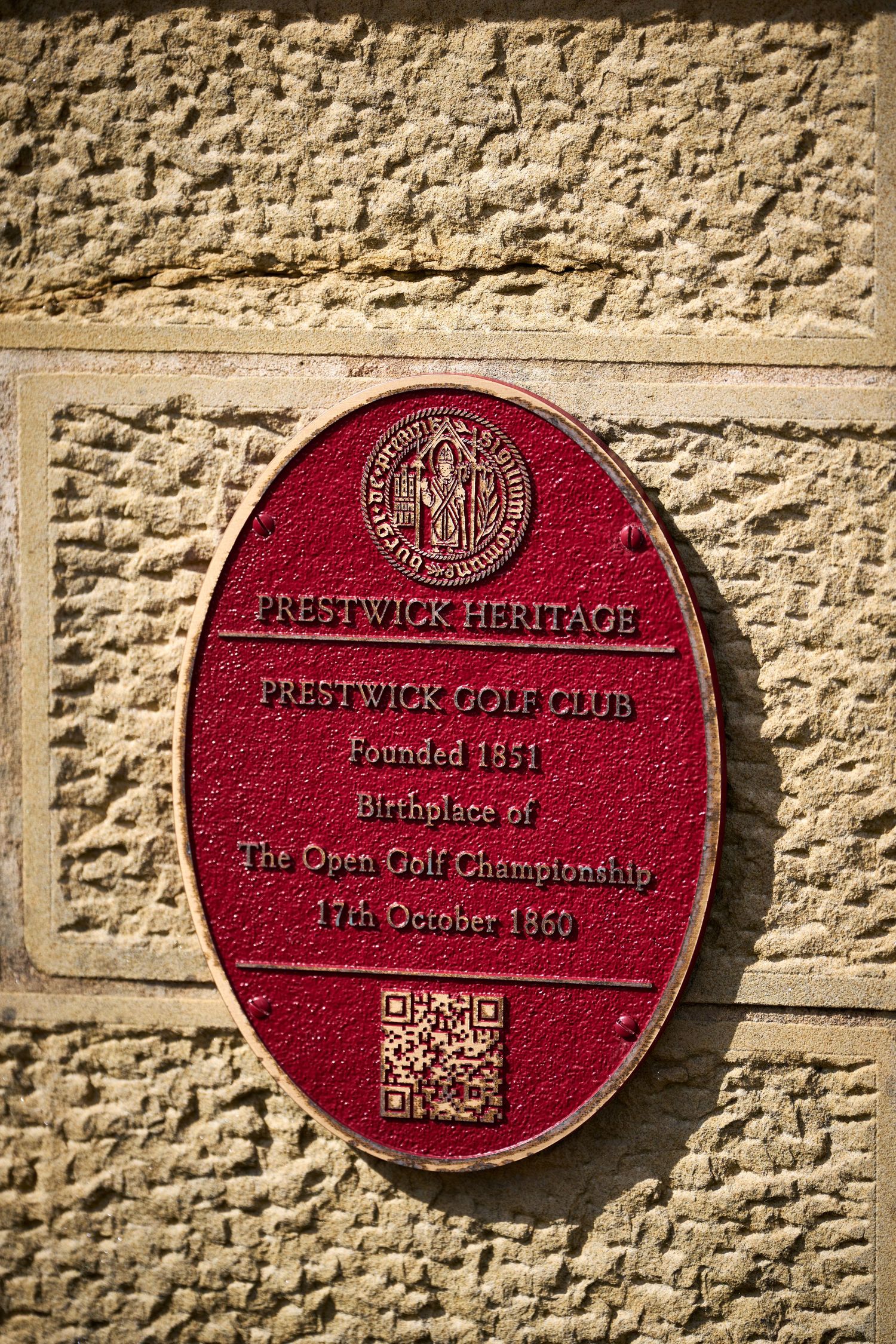 Red heritage plaque for Prestwick Golf Club, founded 1851, birthplace of The Open Golf Championship, mounted on stone wall.
