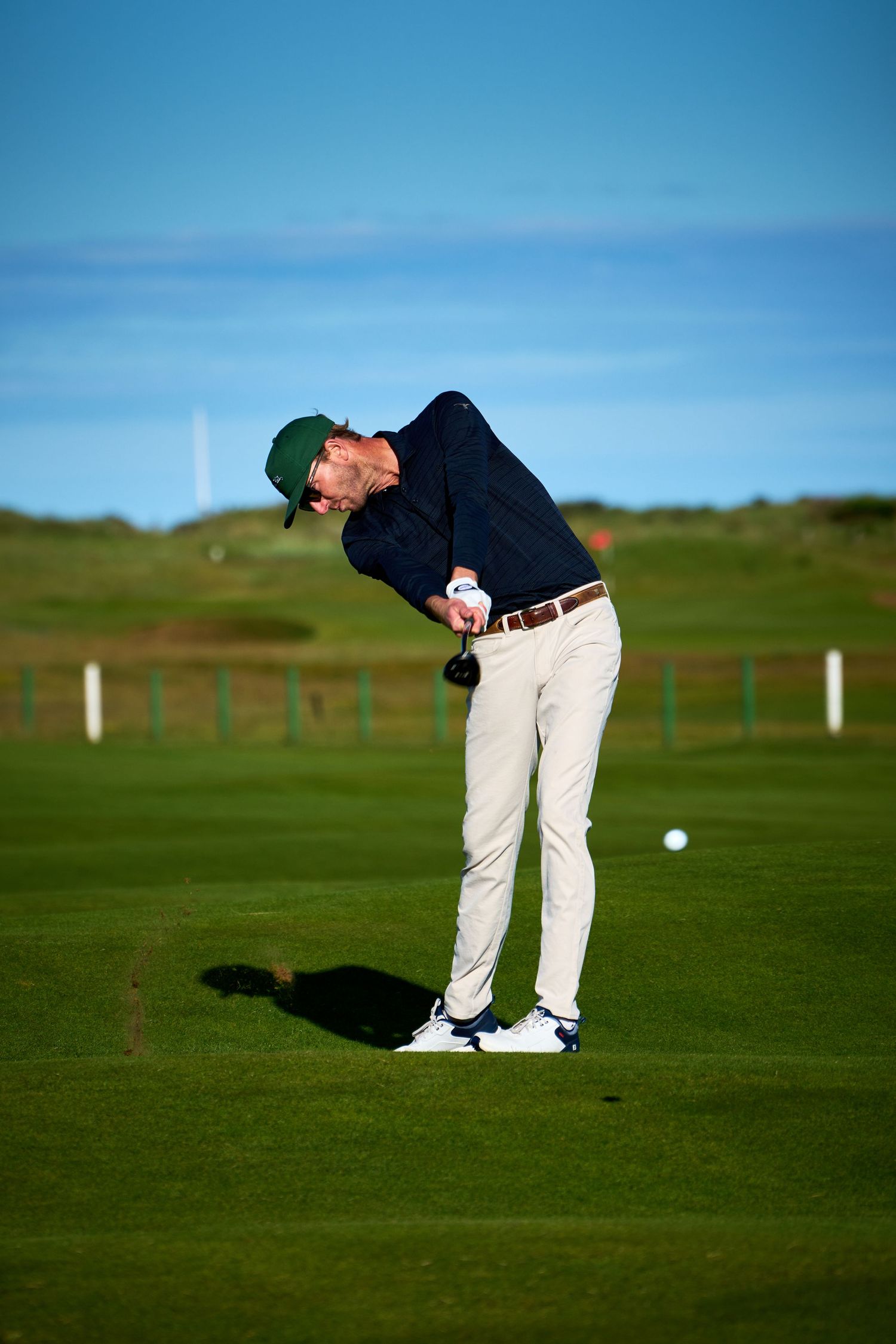 Golfer in navy sweater and khaki pants mid-swing on a lush green golf course under a clear blue sky.