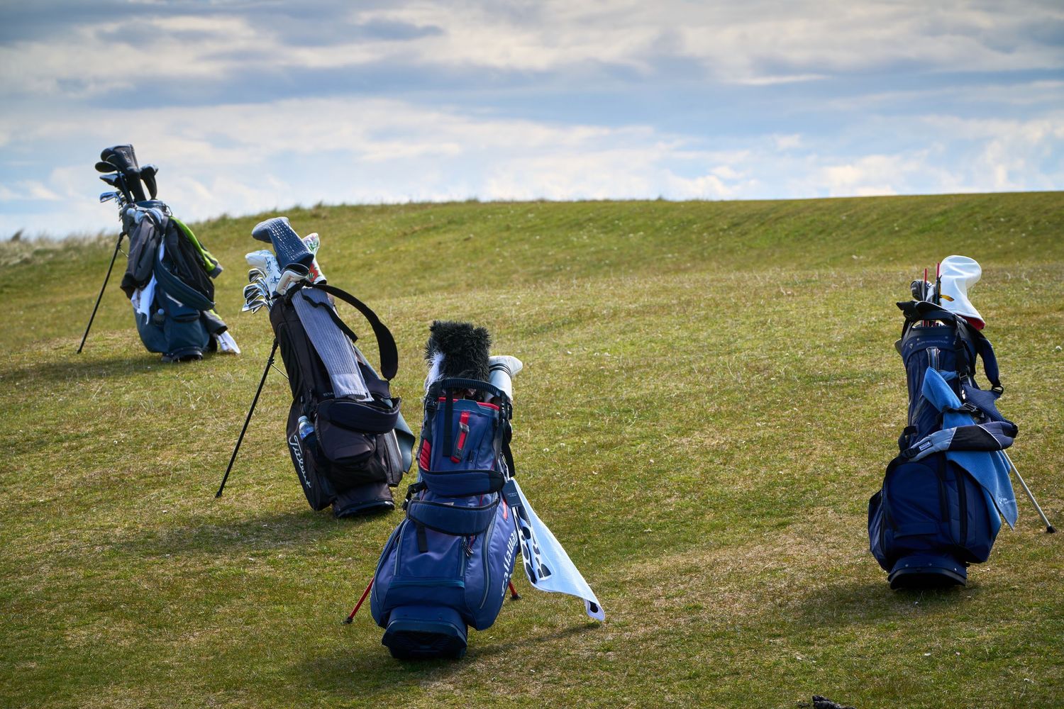 Three blue golf bags standing upright on a green hillside fairway under a partly cloudy sky.