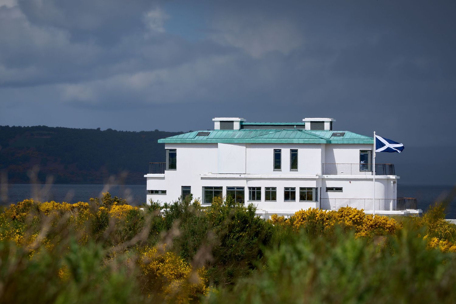 White coastal building with green roof flying Scottish flag, surrounded by yellow gorse bushes overlooking a bay.