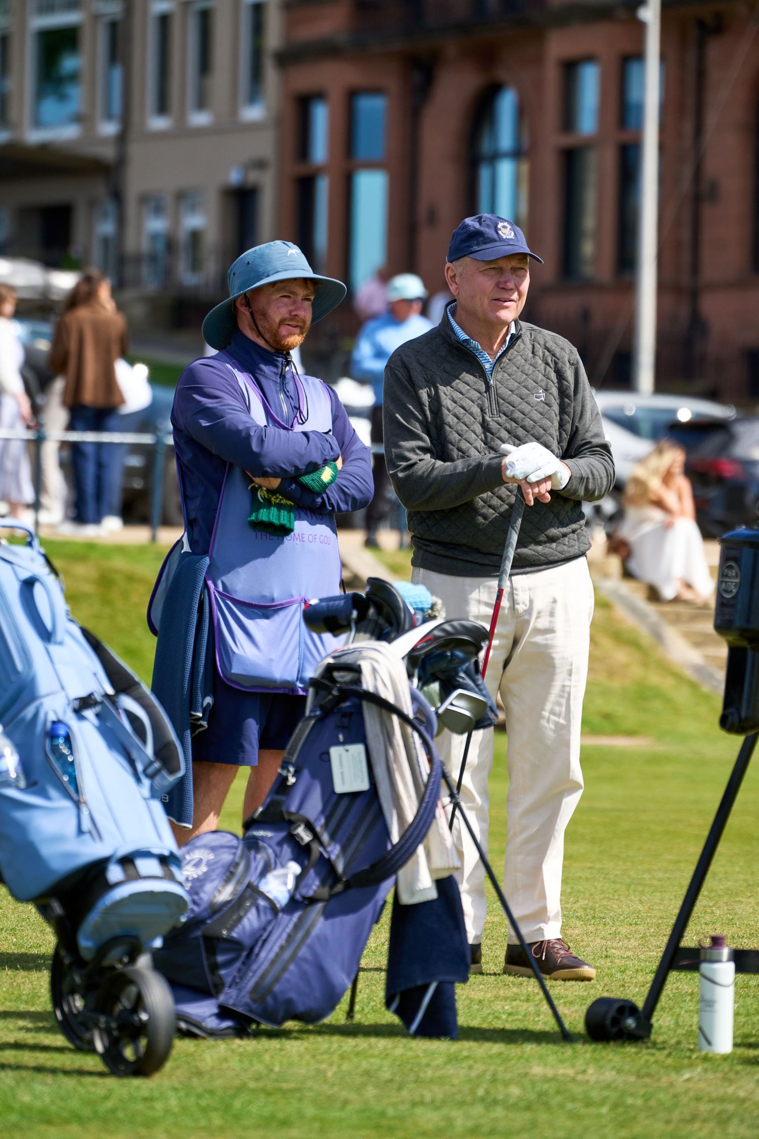 Two golfers stand beside blue golf bags on a sunny course near a brick clubhouse.