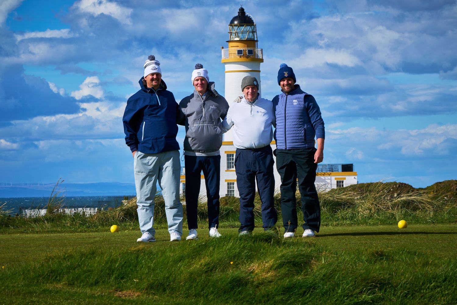 Four golfers pose together on a golf course with a historic lighthouse backdrop under a cloudy sky.