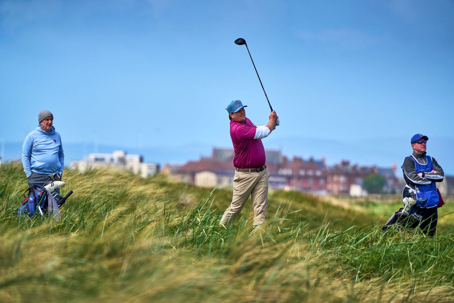 Golfer mid-swing on a links course with caddies watching, tall grass and coastal town in the background.