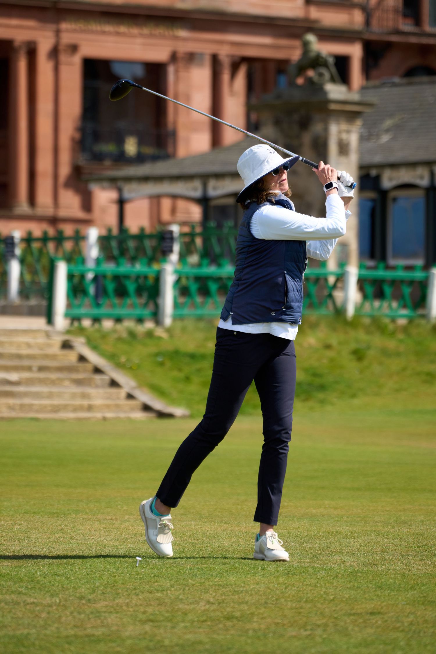Woman golfer mid-swing on a sunny golf course, wearing navy vest and white hat, with a red brick clubhouse behind.
