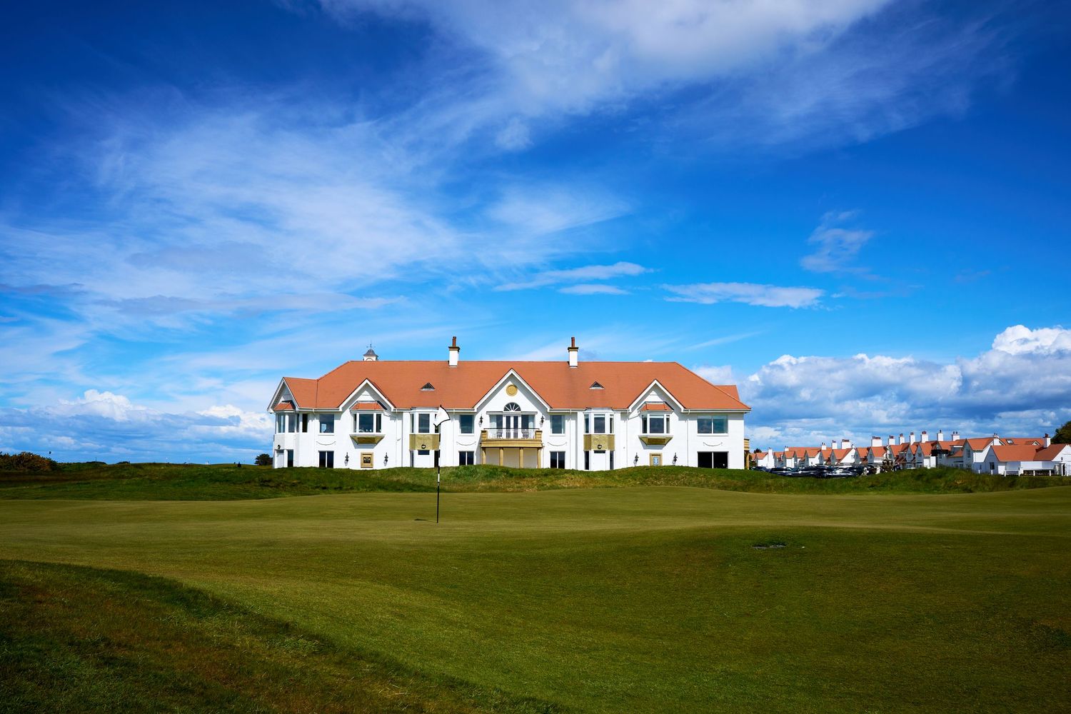 A scenic golf course with lush green fairways and a classic white clubhouse under a bright blue sky.
