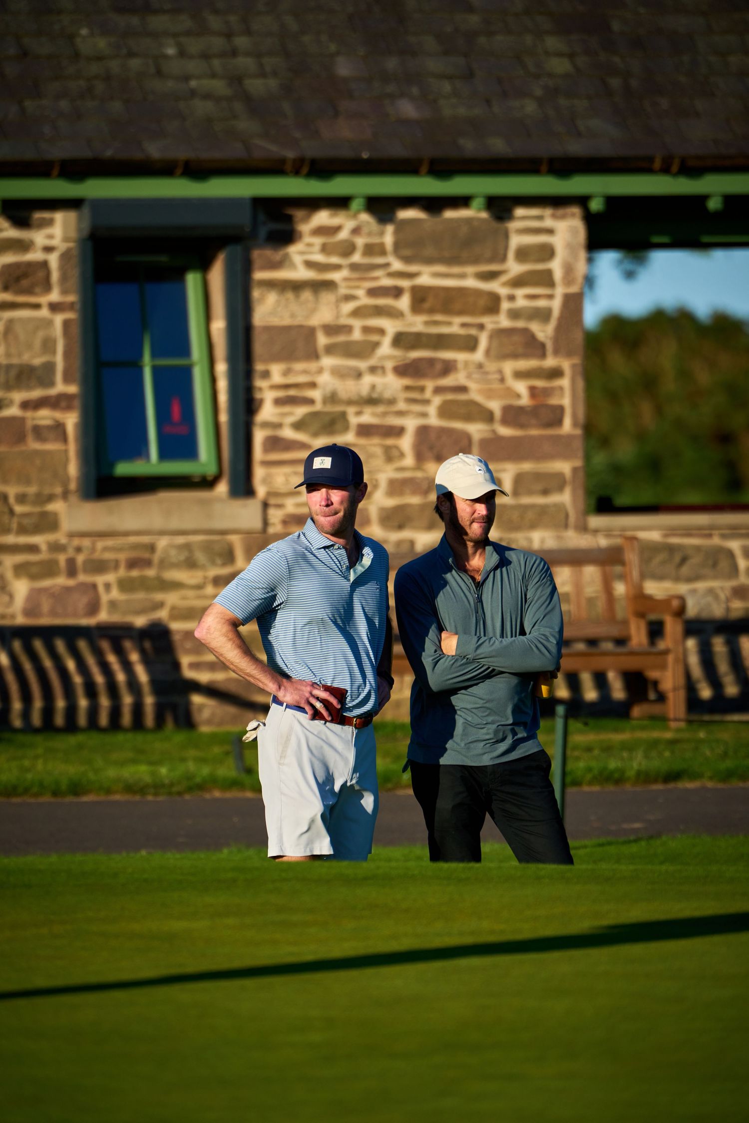Two male golfers stand on a sunny golf course green in front of a rustic stone building.
