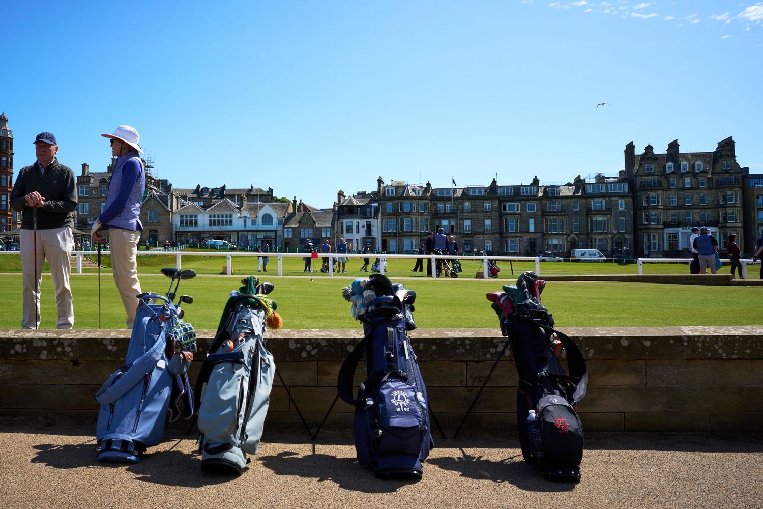 Golf bags lined up on a bridge at St Andrews Old Course with the historic clubhouse in background.