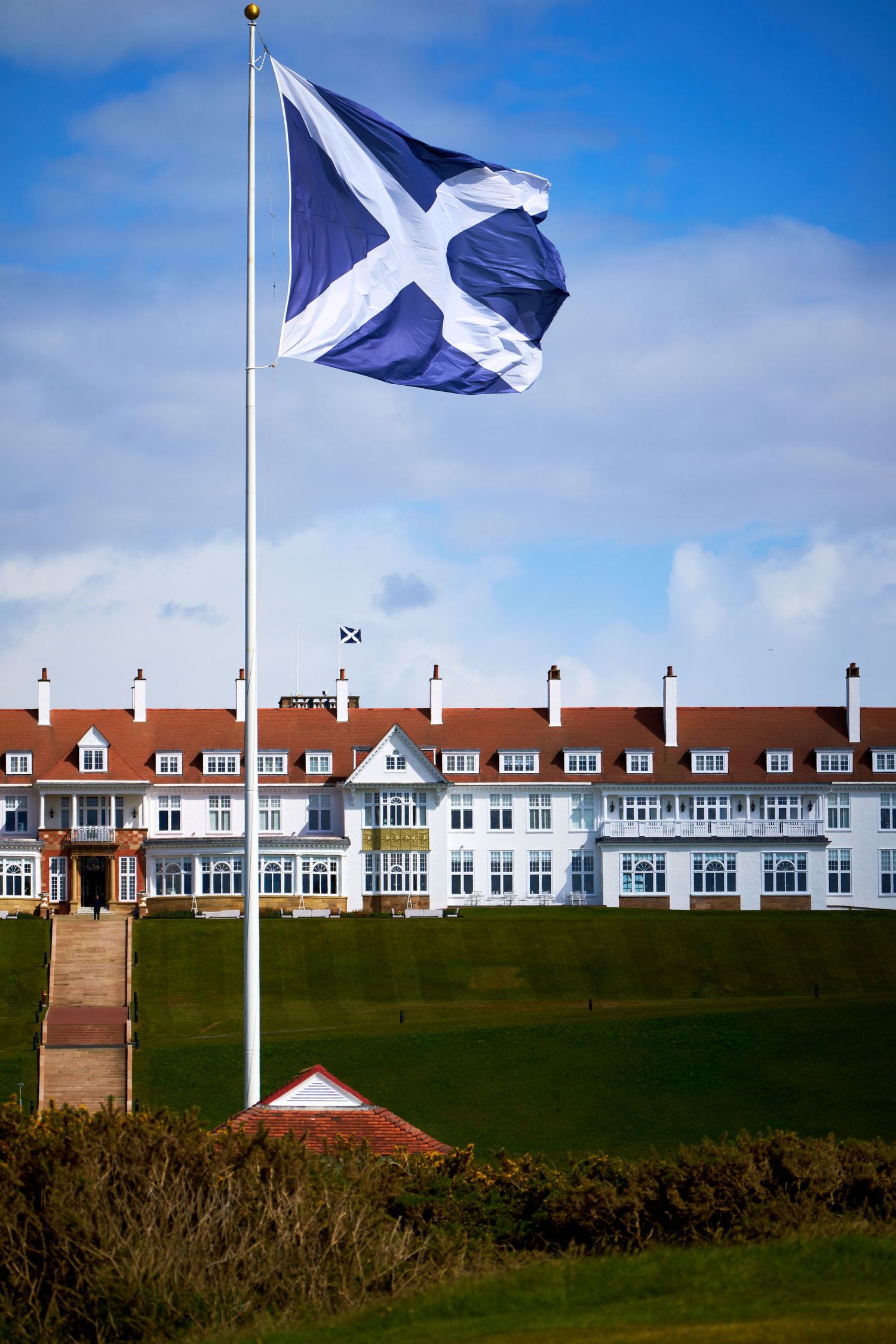 Scottish Saltire flag flying proudly on a tall flagpole in front of a grand white hotel building under a blue sky.