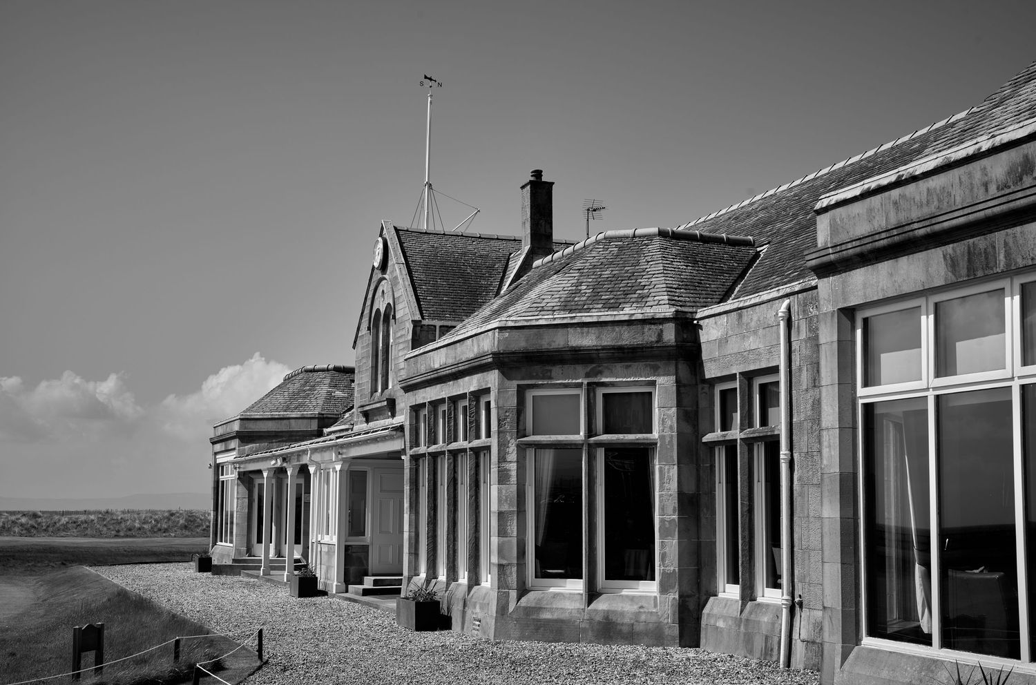 Black and white photo of a historic stone building with large windows beside a coastal beach landscape.