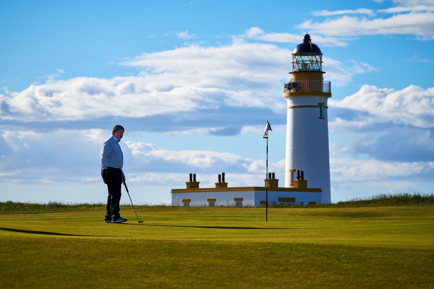 Golfer putting on a green beside a white lighthouse under a blue sky with clouds.