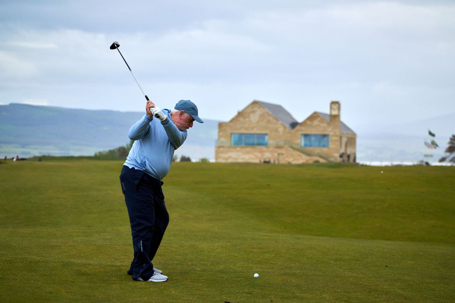 Golfer swinging a club on a scenic coastal golf course with a stone clubhouse in the background.