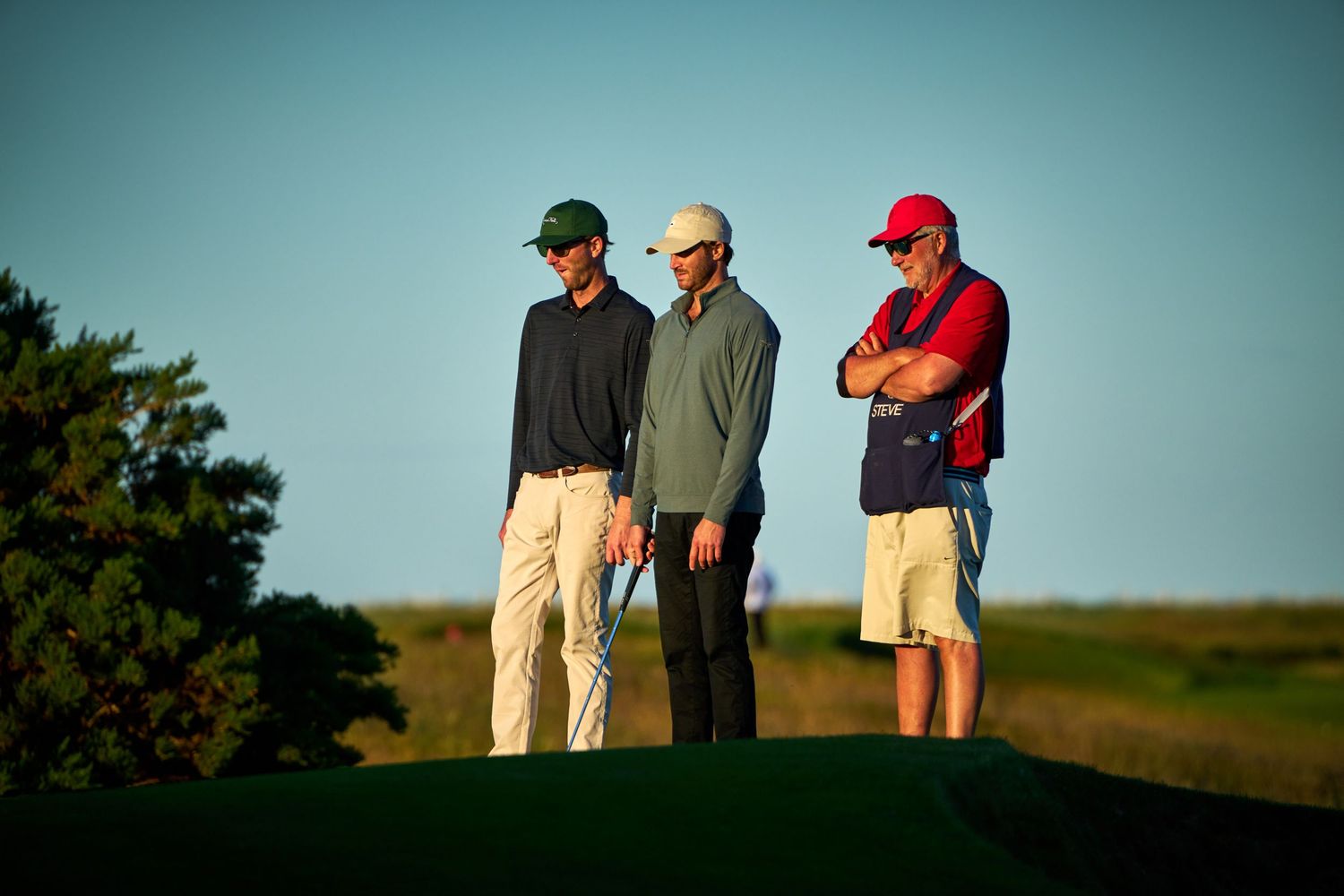 Three golfers standing on a golf course at golden hour, wearing casual golf attire including hats.