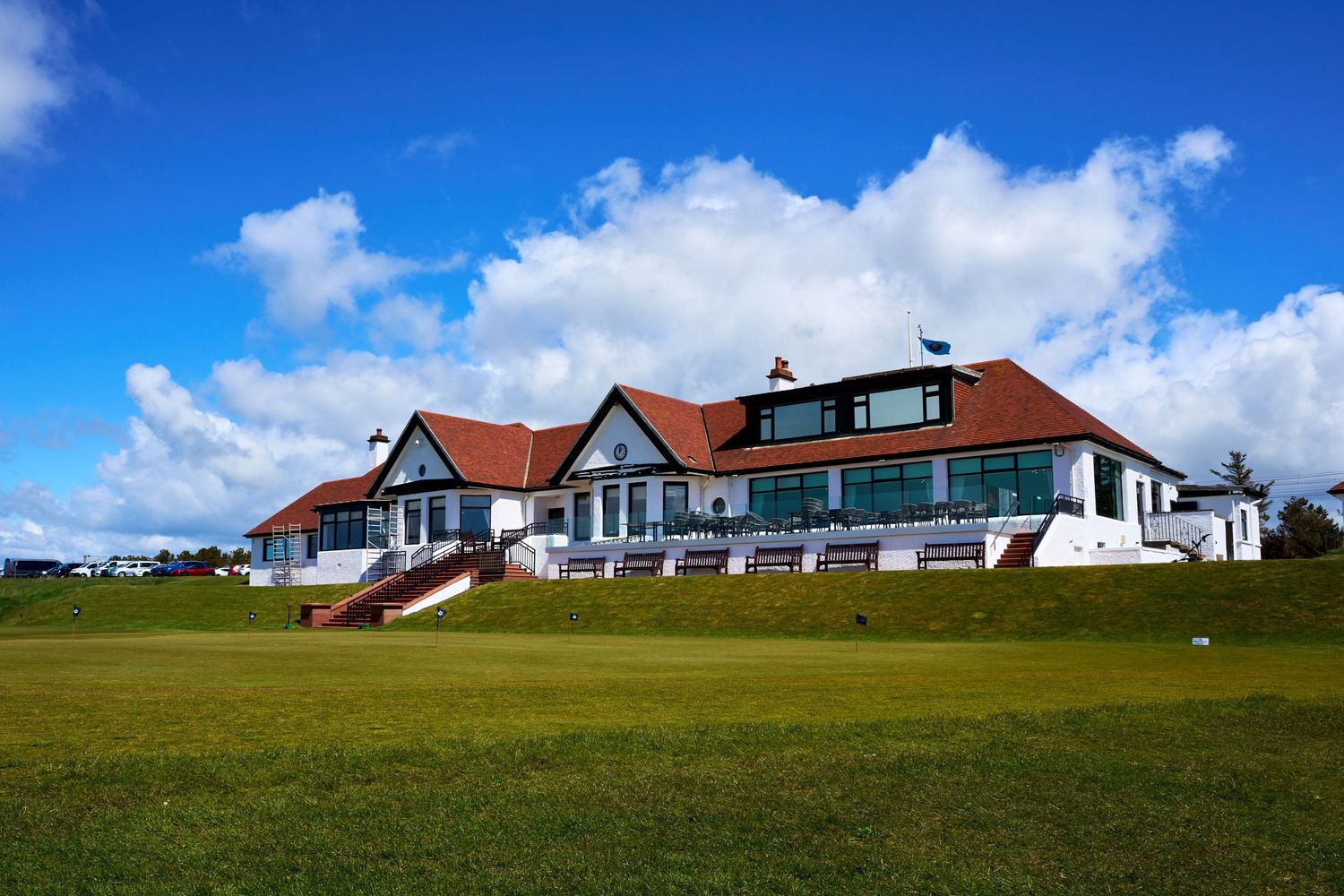 Golf clubhouse with red roof and white facade overlooking lush green fairway under bright blue sky with clouds.