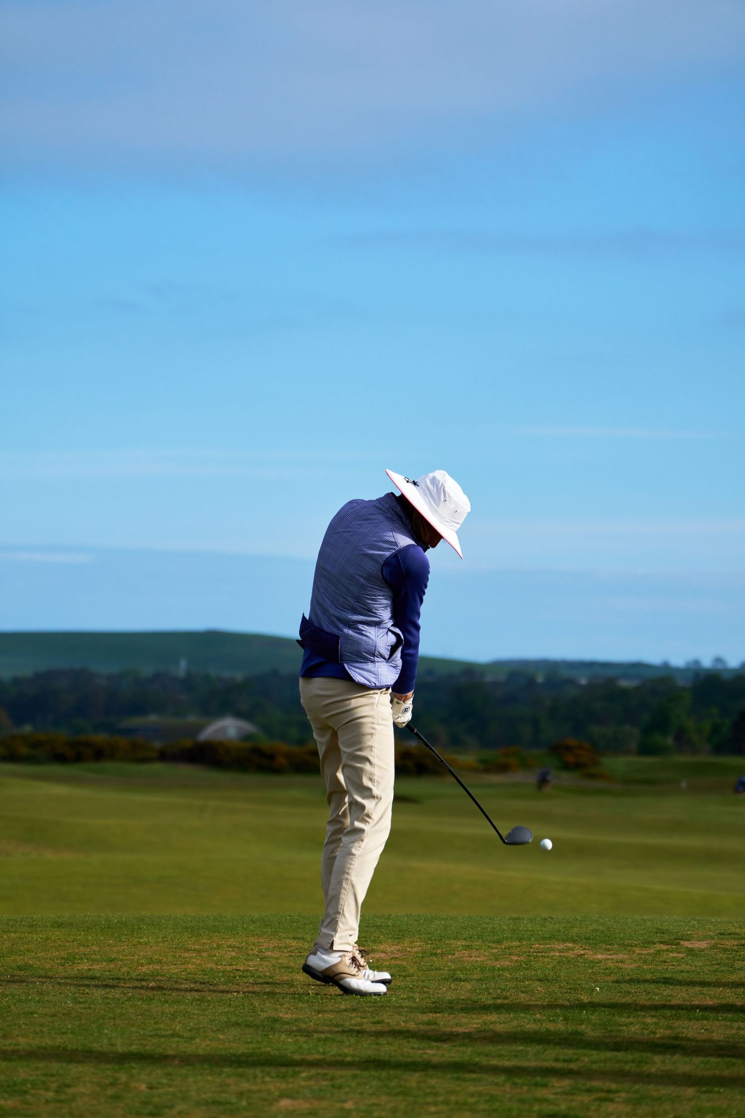 Golfer in white hat and blue shirt swinging a club on a sunny green golf course with blue skies.