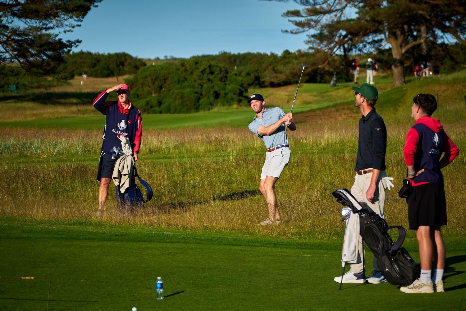 Golfers on a scenic course, one mid-swing while others watch, with golf bags and lush green fairways.