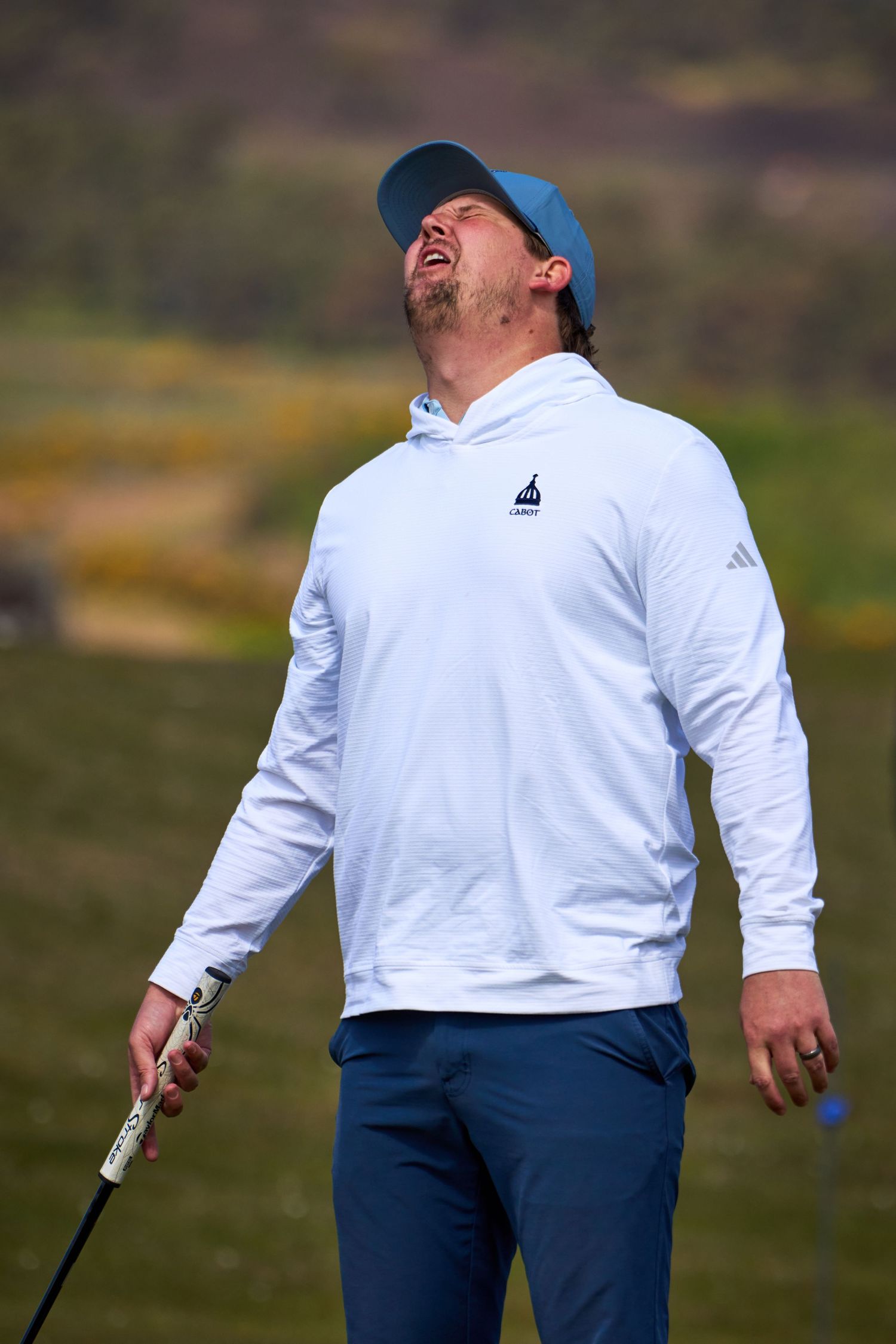 Frustrated golfer in white Adidas hoodie and blue cap reacts after a missed shot on a golf course.