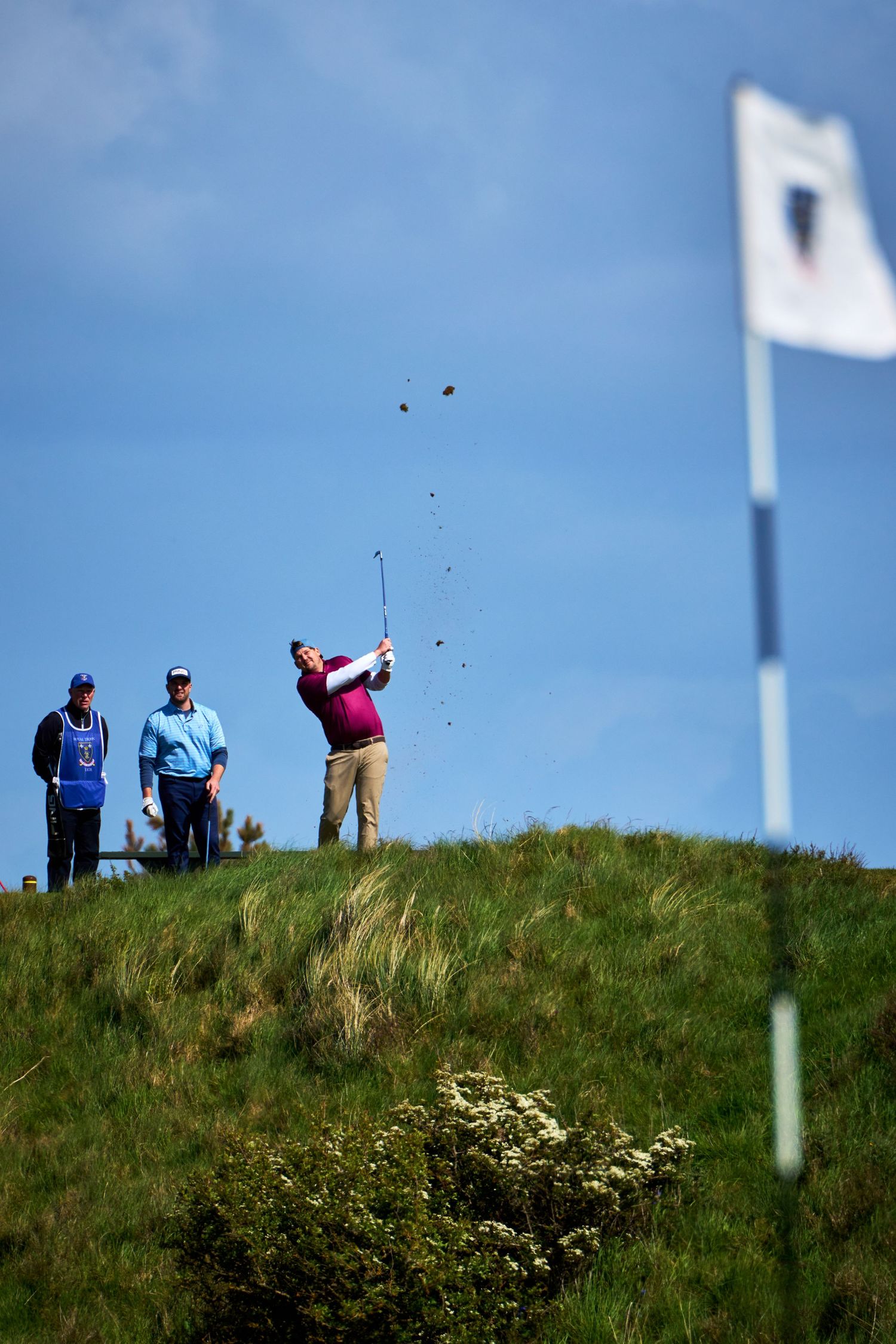 Golfer in pink shirt hits a shot from rough hillside as caddie and spectator watch, white flag visible against blue sky.