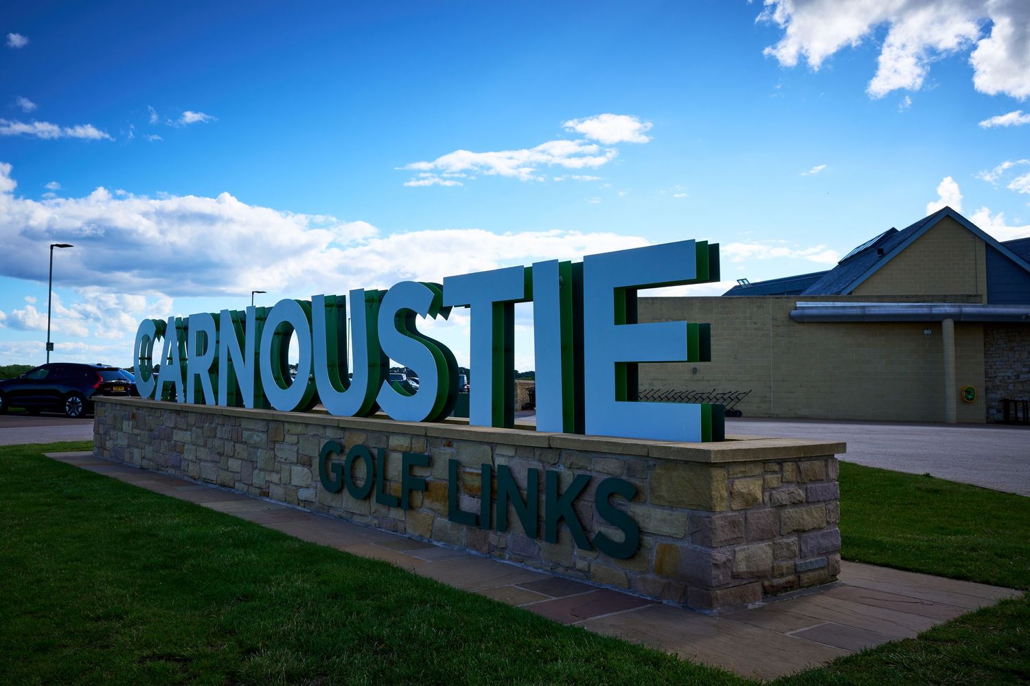 Carnoustie Golf Links sign mounted on a stone wall under a blue sky with clouds in the background.