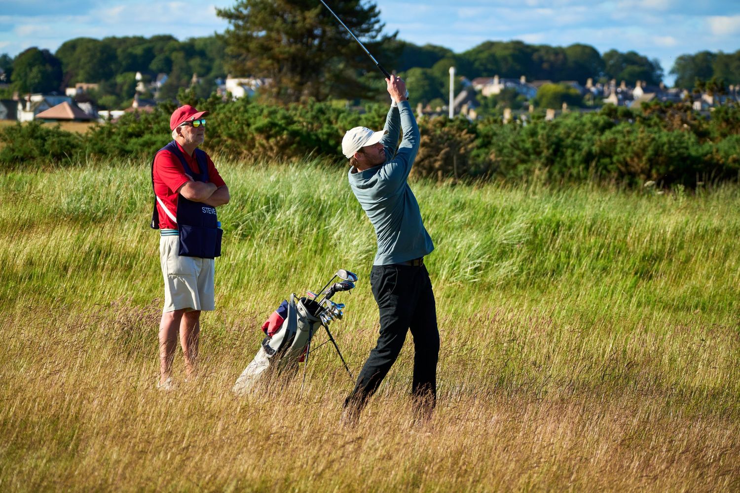 Golfer swinging a club in tall grass rough while a caddie watches, with a golf bag nearby on a sunny day.