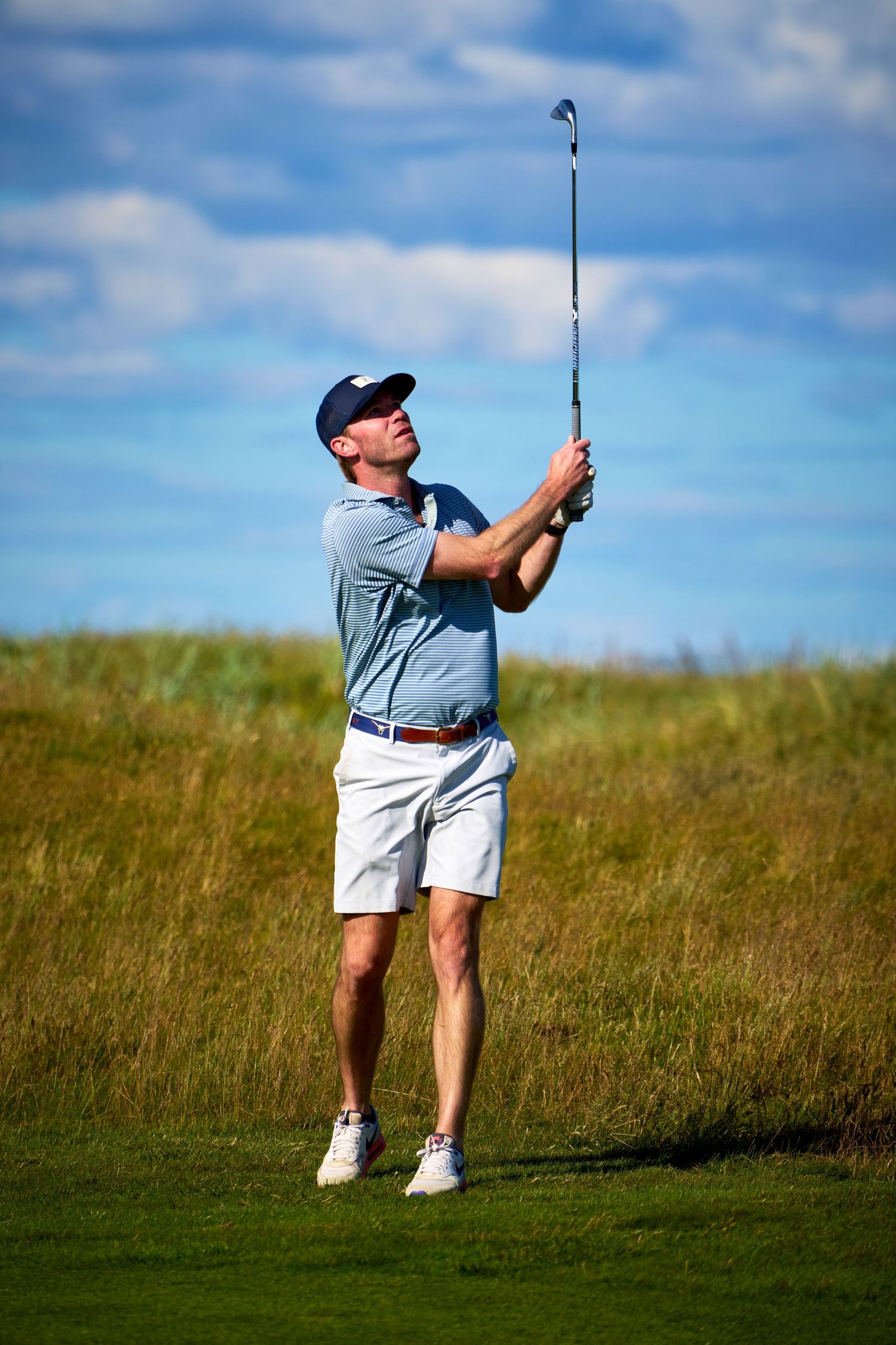 Male golfer in white shorts and blue shirt follows through on a swing on a sunny golf course.