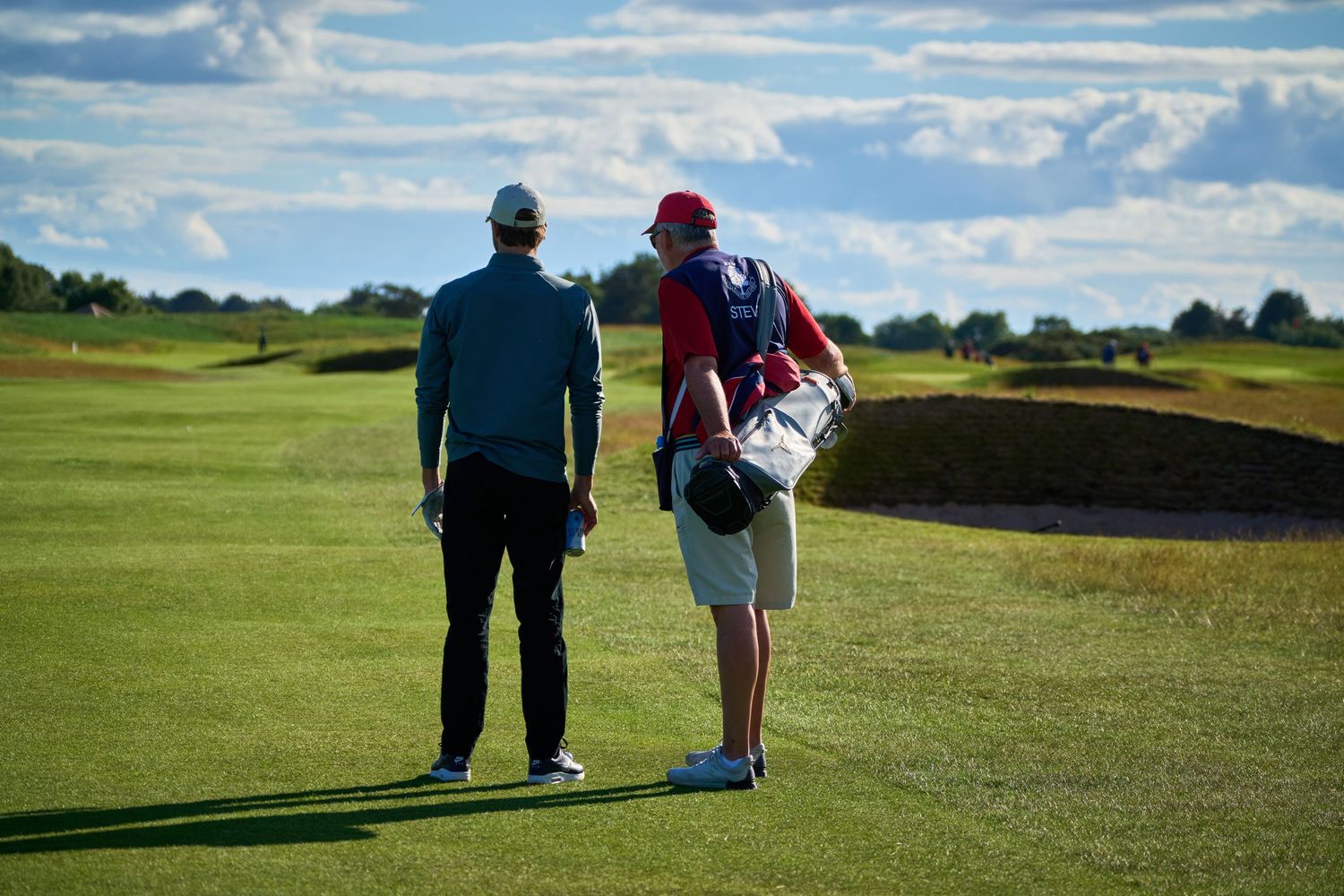 Two golfers walk along a lush green fairway, one carrying a golf bag, under a partly cloudy sky.