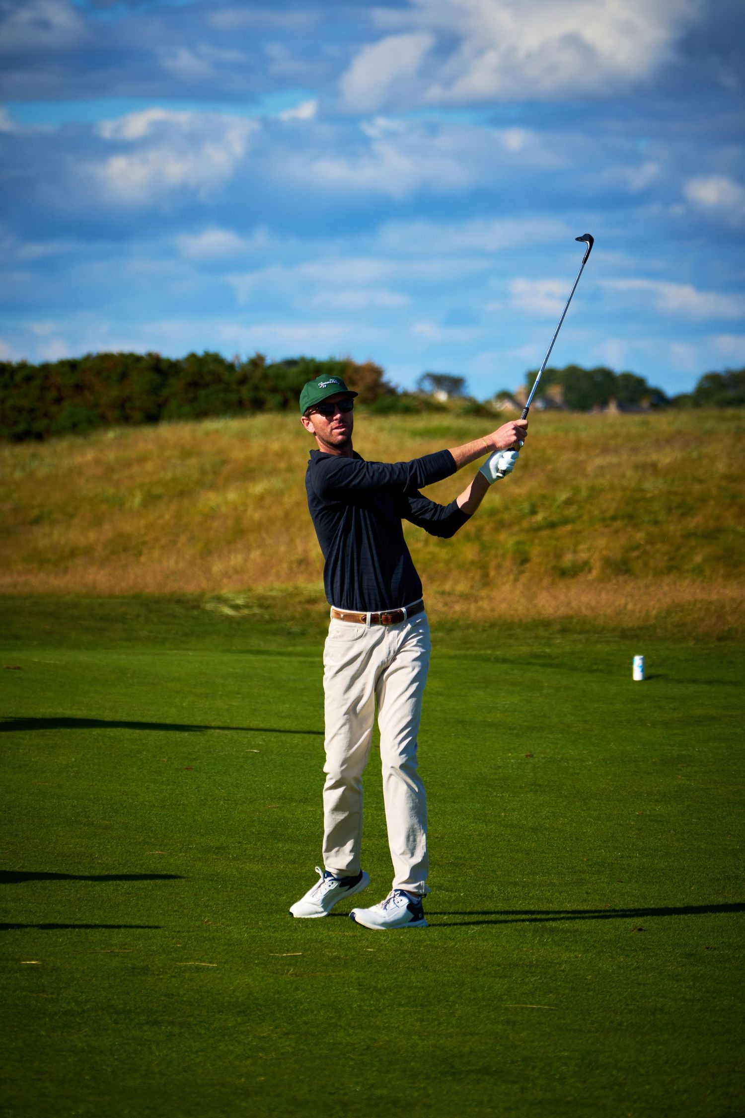 Golfer in navy shirt and white pants completing a swing on a sunny golf course with blue skies.