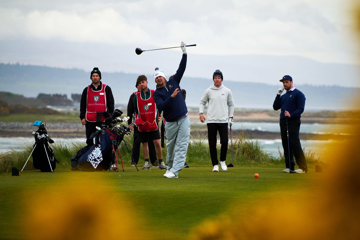 Golfer swinging club on scenic coastal golf course with caddie and spectators watching on a cloudy day.