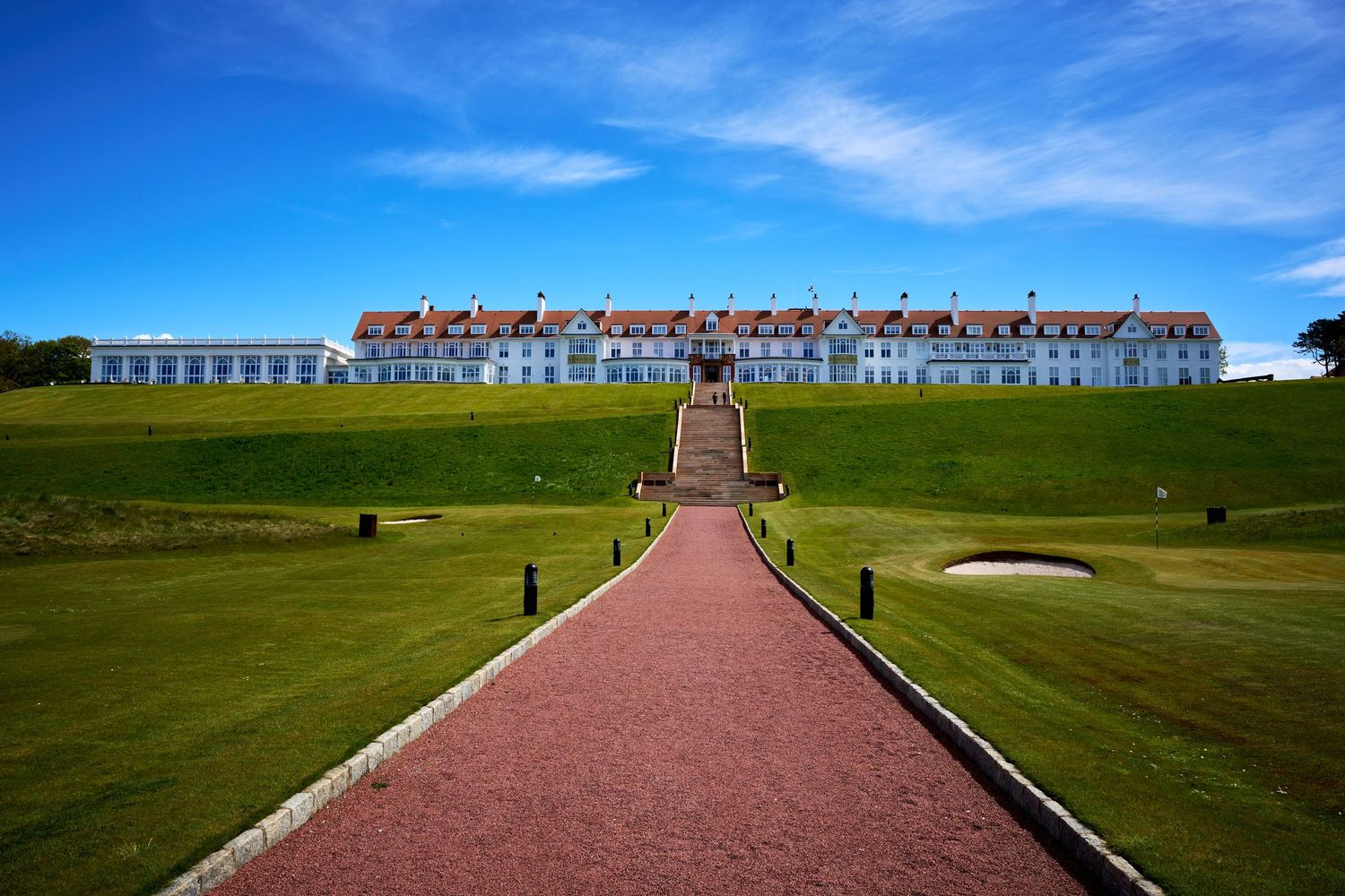 Grand white hotel building overlooking a lush green golf course under a bright blue sky with a red brick pathway leading to the entrance.