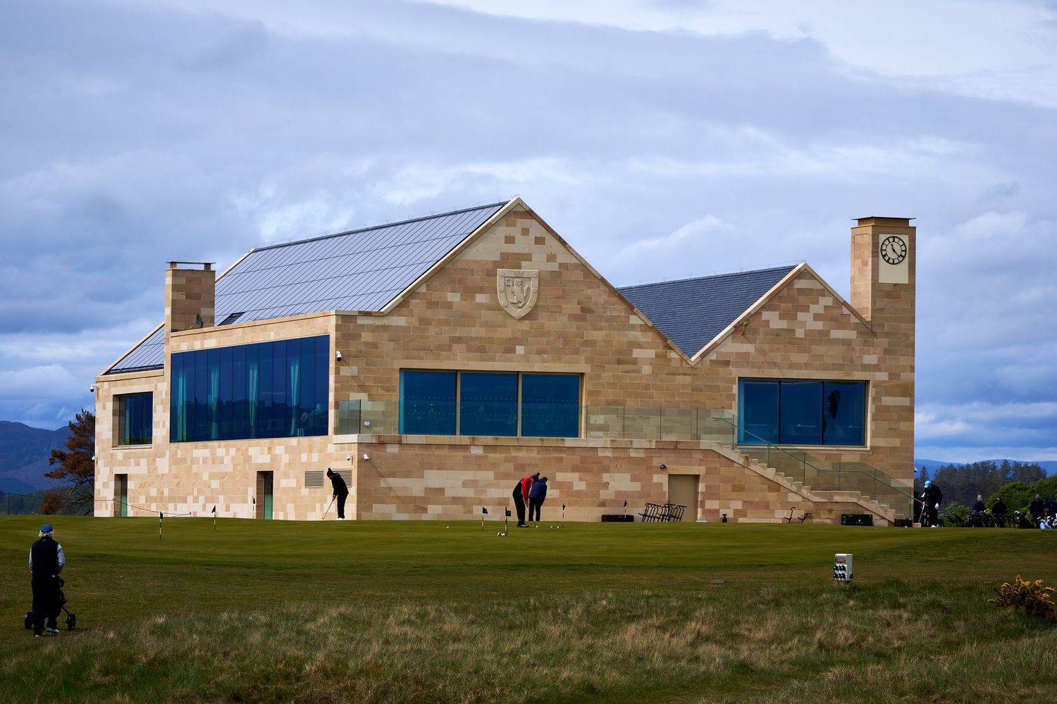 Modern stone golf clubhouse with clock tower, solar panels, and large windows on a lush green course under cloudy skies.