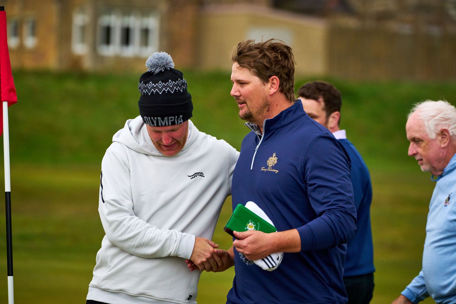 Two golfers shaking hands on a golf course during a tournament, wearing casual sports attire.