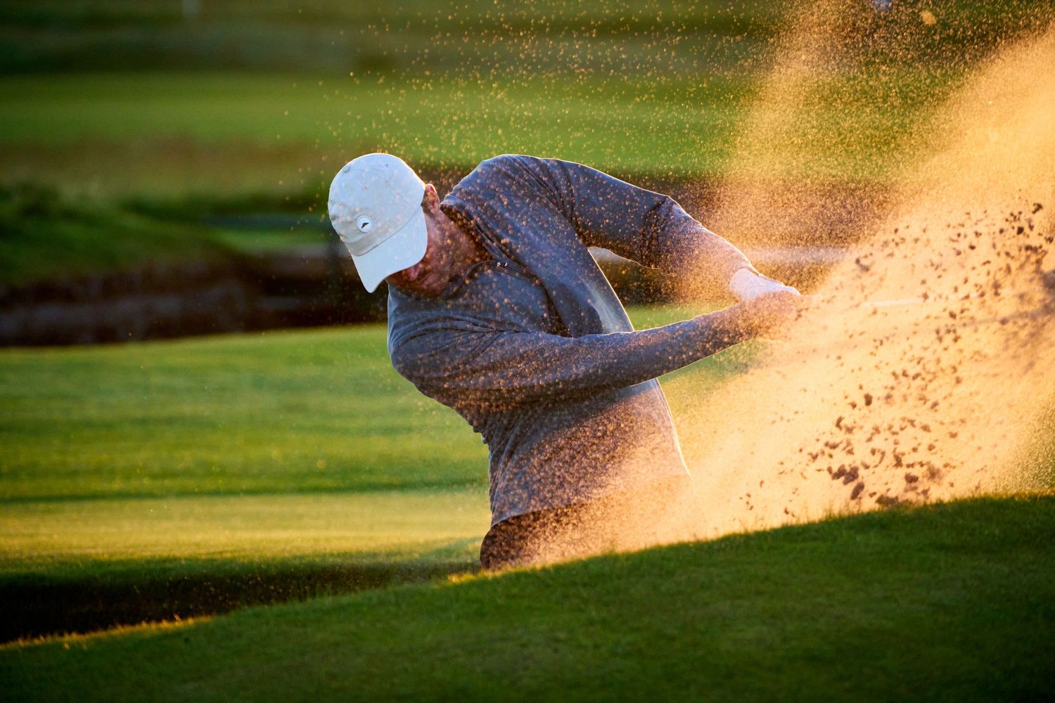 Golfer hitting a shot from a sand bunker, sending a spray of sand into the air on a sunlit golf course.