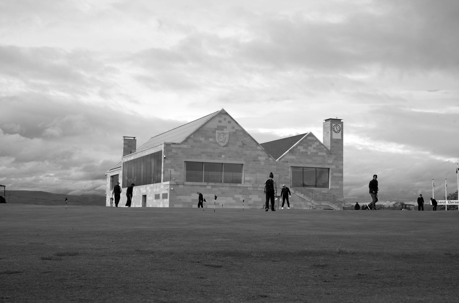 Black and white photo of a stone building on an open field with people walking nearby under a cloudy sky.