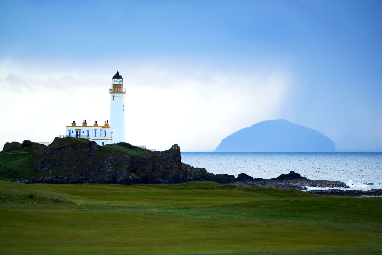 A white lighthouse stands on rocky Scottish coastline with lush green golf course and Ailsa Craig island in the background.
