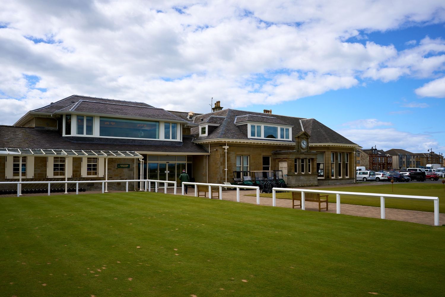 Historic brick clubhouse with white railings and pergola beside a manicured green lawn under a partly cloudy sky.