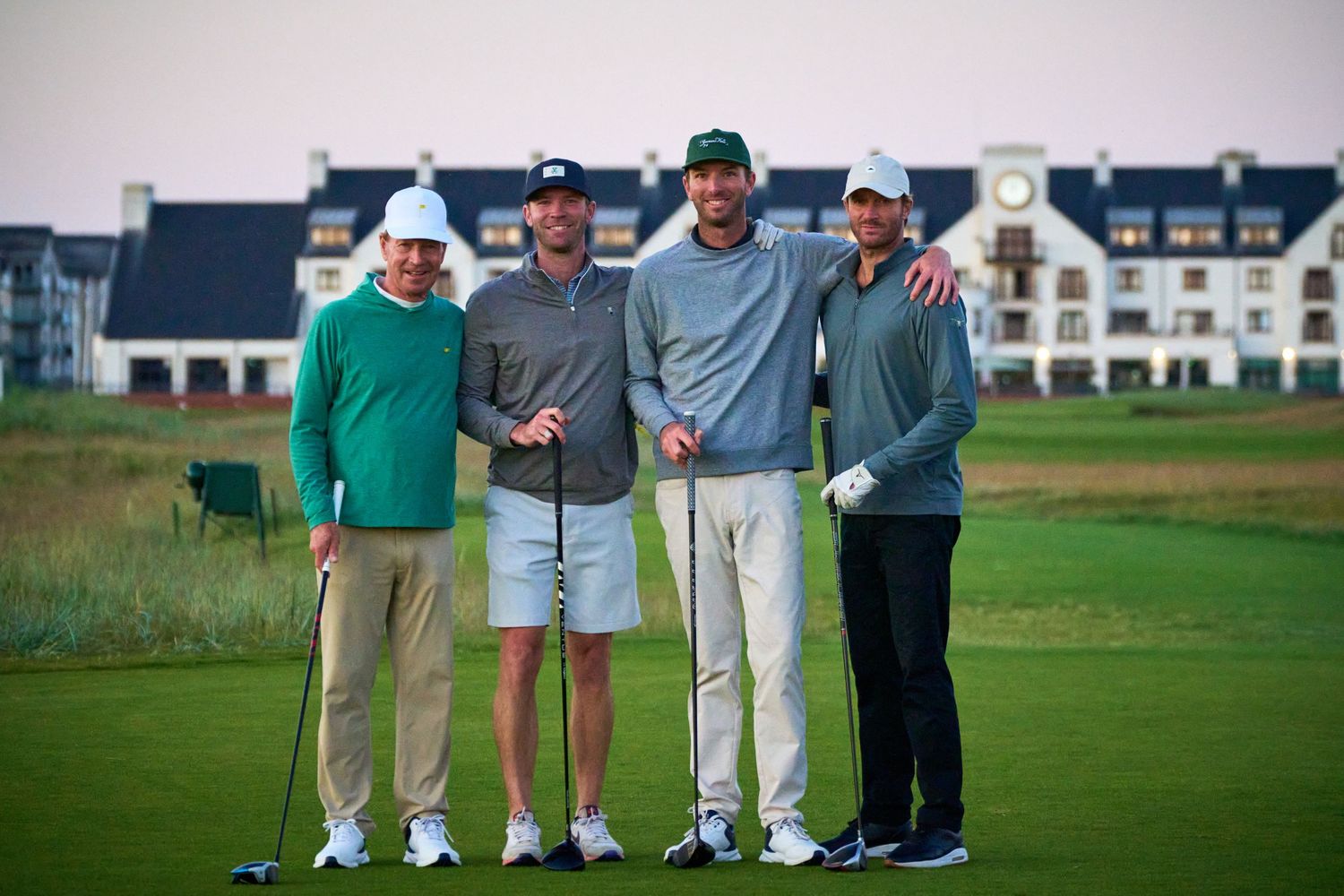 Four men posing together on a golf course with a large resort building in the background at dusk.