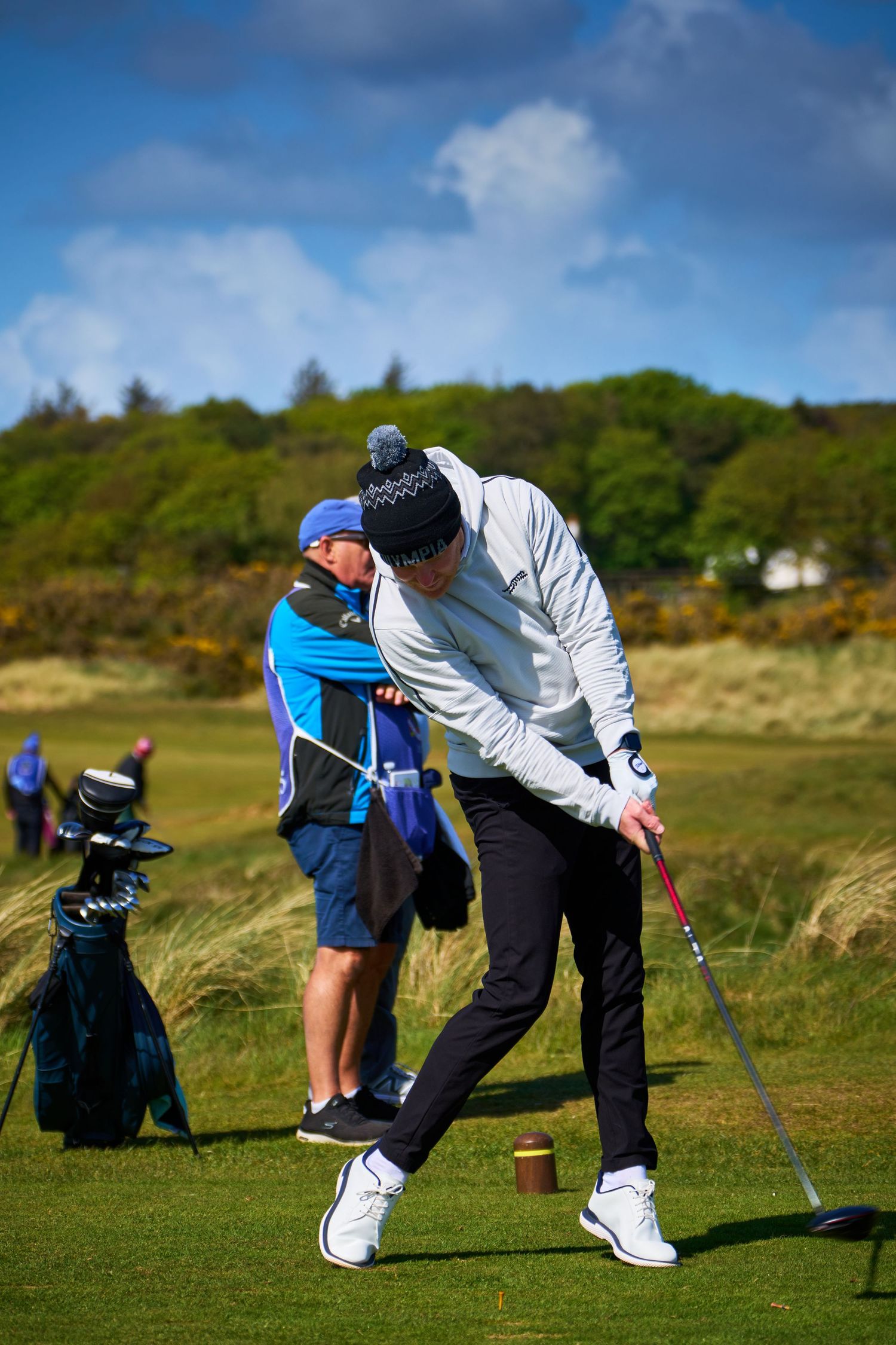 Golfer mid-swing on a links course with caddie watching, wearing winter hat and white jacket on a sunny day.