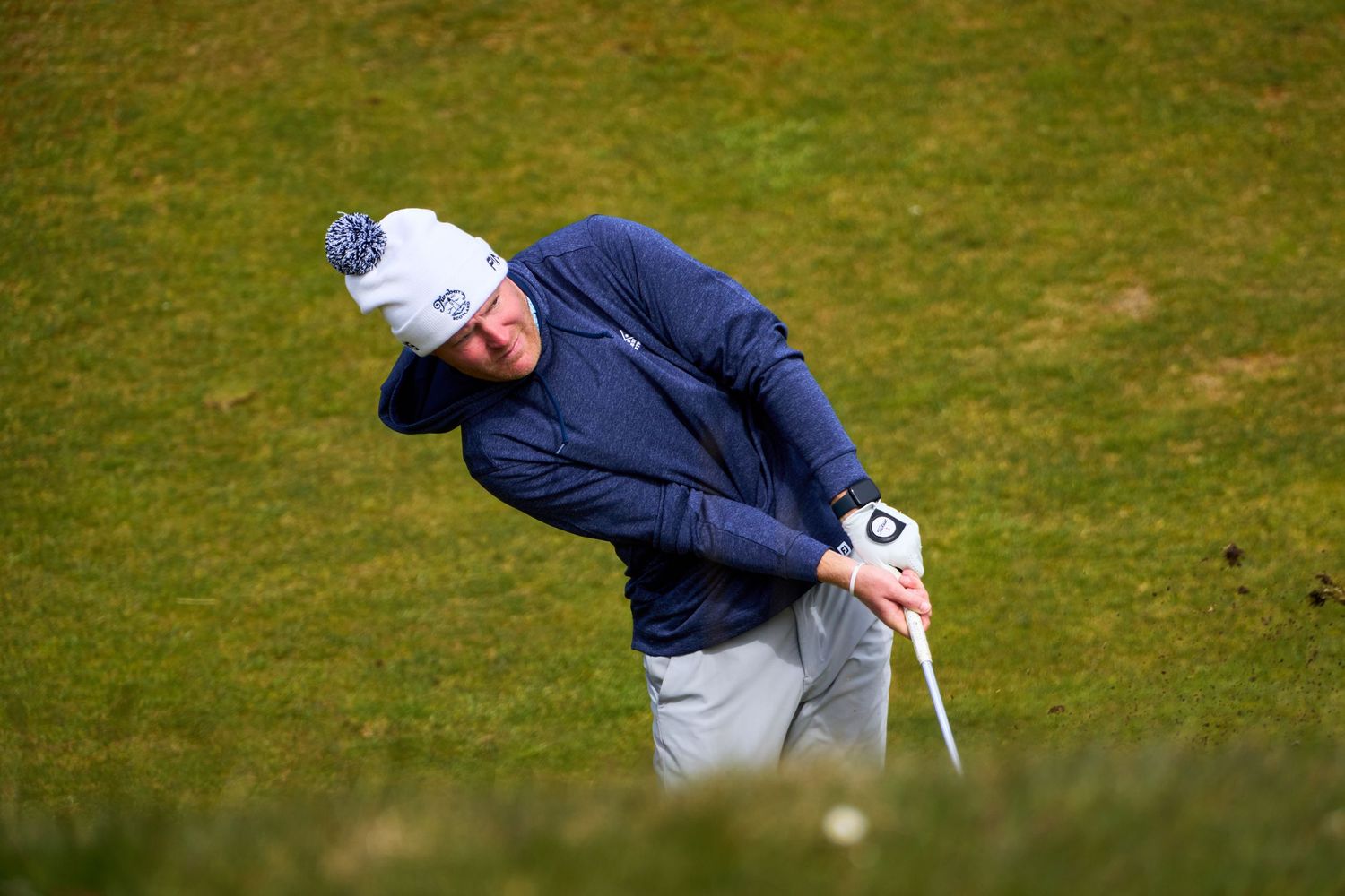 Golfer in navy jacket and white beanie executing a chip shot on a links golf course.