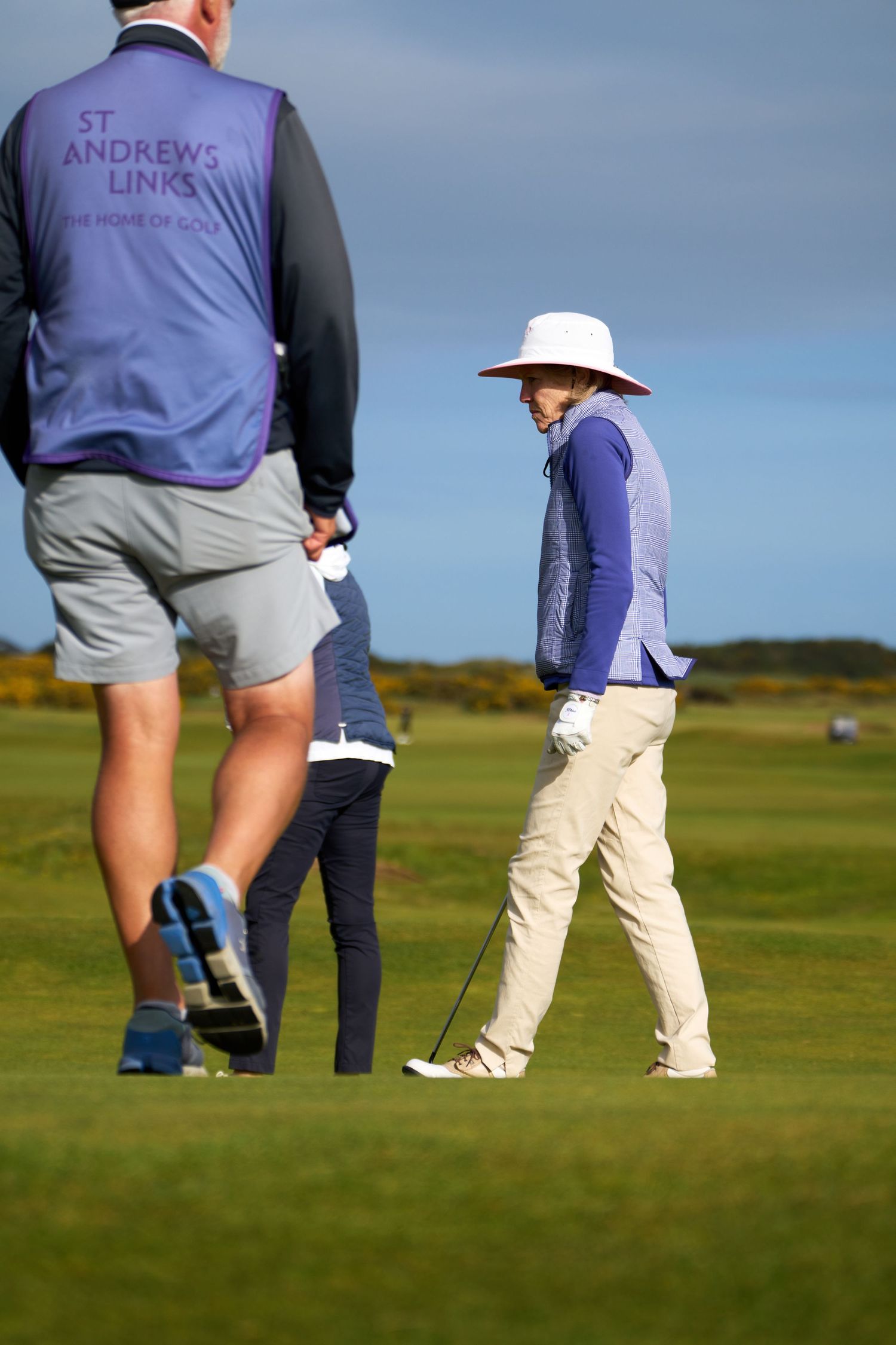 Golfer in white hat and blue vest walks the fairway at St Andrews Links, the Home of Golf, on a sunny day.
