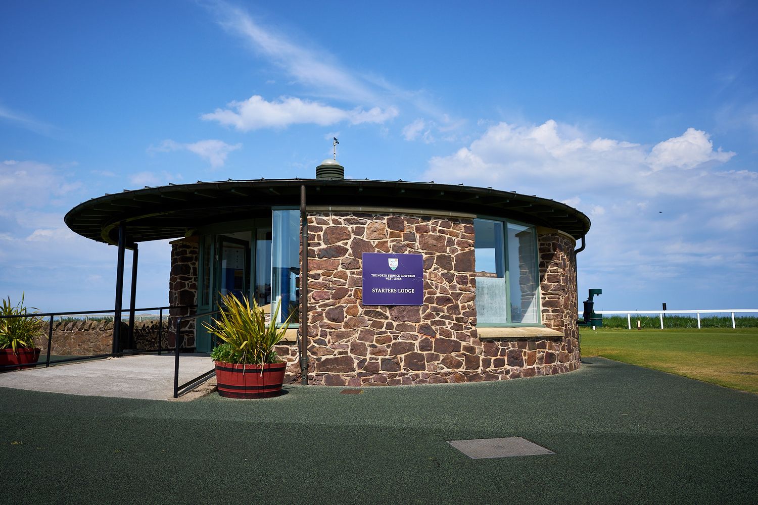 Circular stone building with blue sign, green lawn, and ocean views under a bright blue sky with white clouds.